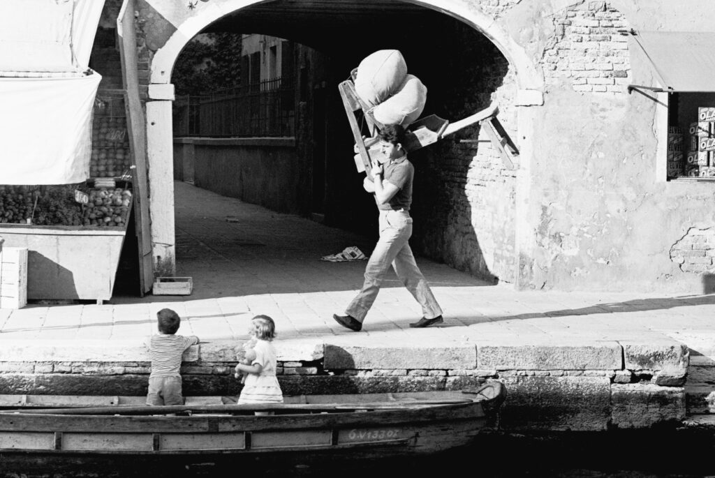 La foto in bianco e nero di un materassaio mentre passa davanti a un sottoportico con un materasso sulle spalle in Fondamenta delle Cappuccine, due bambini appoggiati al rio mentre sono su un barchino lo guardano passare
